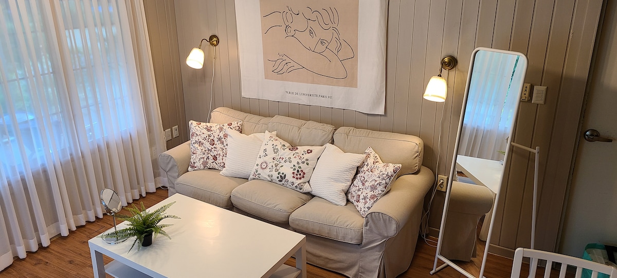 A cozy living area features a light-colored sofa adorned with decorative pillows. A stylish wall art piece hangs above the sofa. Soft lighting is provided by two wall sconces, while a small table is set in front, complemented by a potted plant.