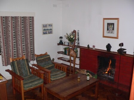 A cozy living area features two wooden chairs, each with patterned upholstery, arranged around a wooden coffee table. A fireplace with a gentle fire flickers in the corner, while decorative shelves display small artworks and books. A patterned curtain adds a touch of warmth.