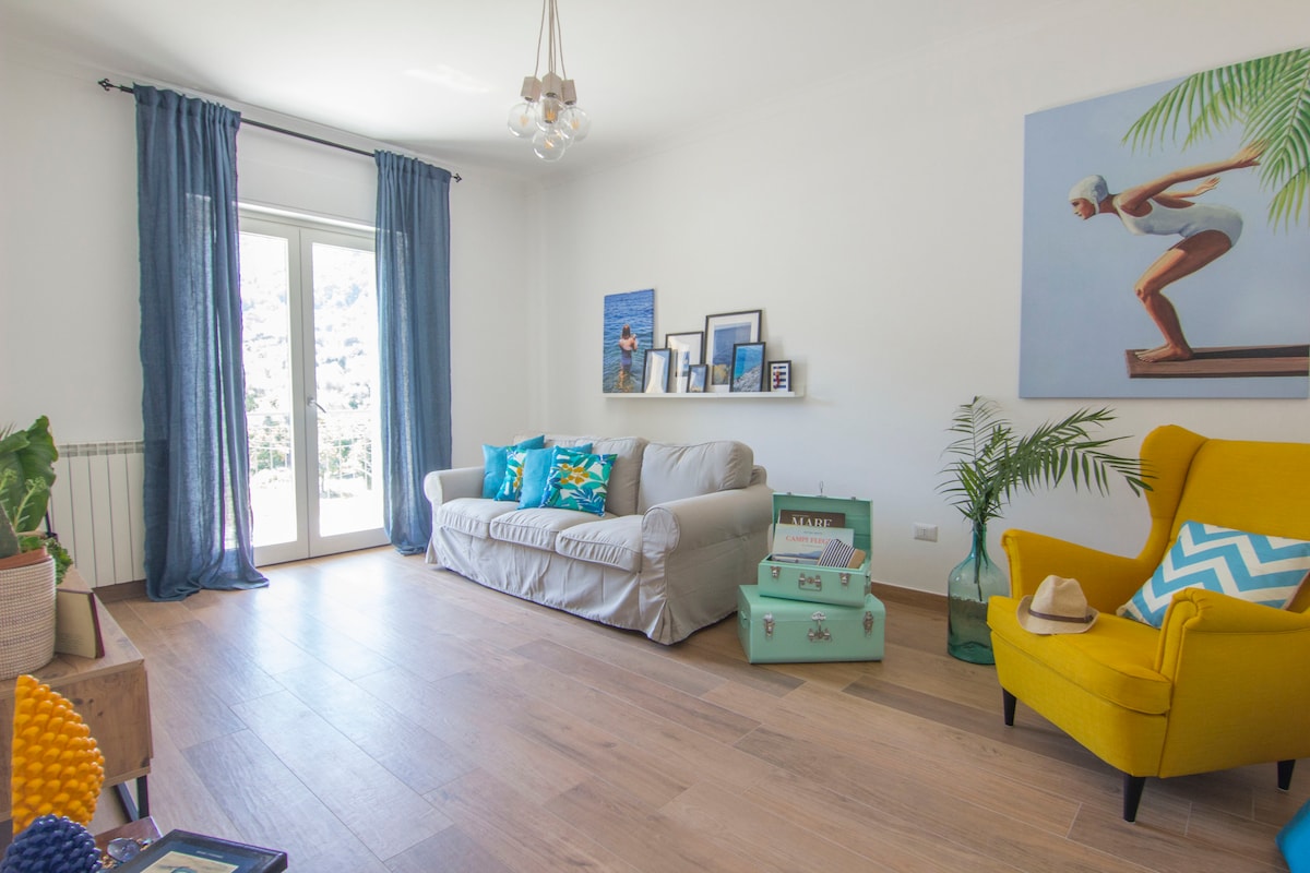 A bright living area features a comfortable light-colored sofa with decorative cushions and a vibrant yellow armchair. A small shelf displays framed photos against a white wall. Large windows allow natural light to fill the space, accompanied by light blue curtains. A potted plant adds a touch of greenery.