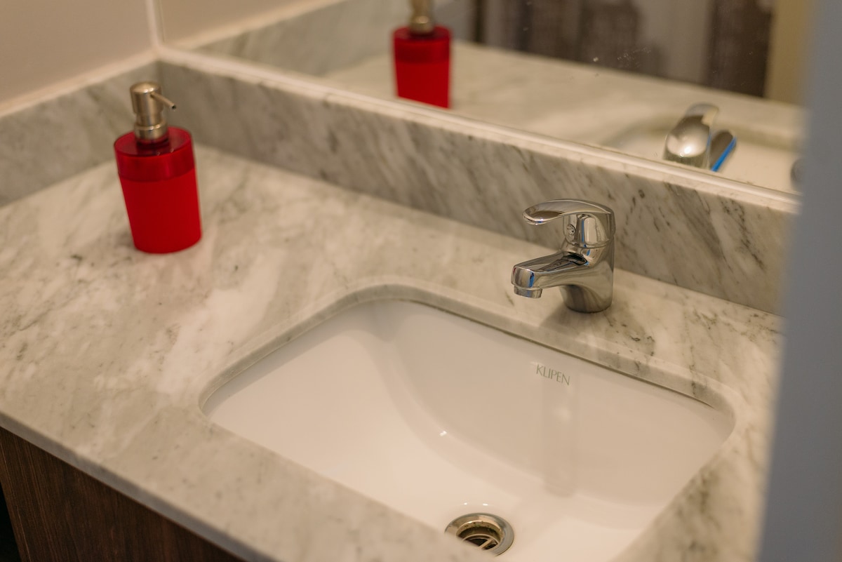 A modern bathroom sink is situated on a marble countertop. A sleek faucet is installed above the white basin, and a red soap dispenser is placed nearby. The mirror above reflects the clean lines of the fixture and countertop.