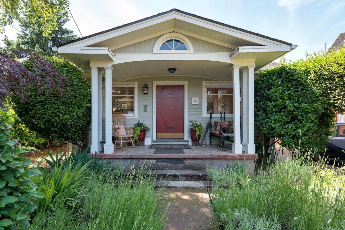 The exterior entrance is framed by lush greenery and flowering plants. A welcoming red door is flanked by two white columns. A small seating area is visible on the porch, with chairs positioned for relaxation. Natural light enhances the inviting ambiance.