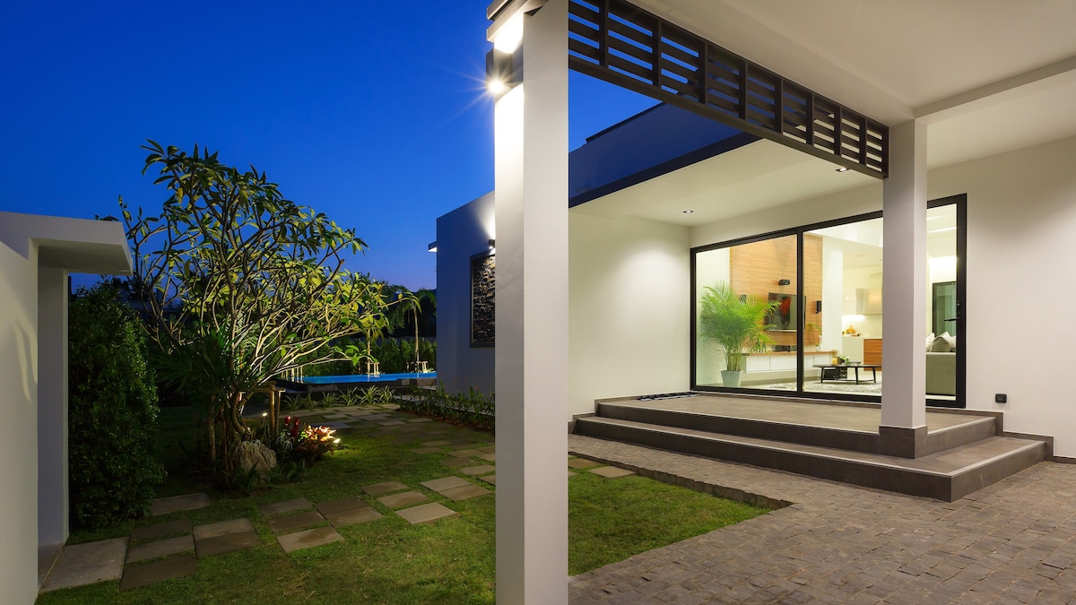 An outdoor view of the villa showcases contemporary architectural elements, with large glass doors revealing the interior. Pebbled pathways lead through a well-maintained garden, featuring some greenery. The serene ambiance is enhanced by soft lighting against the evening sky.
