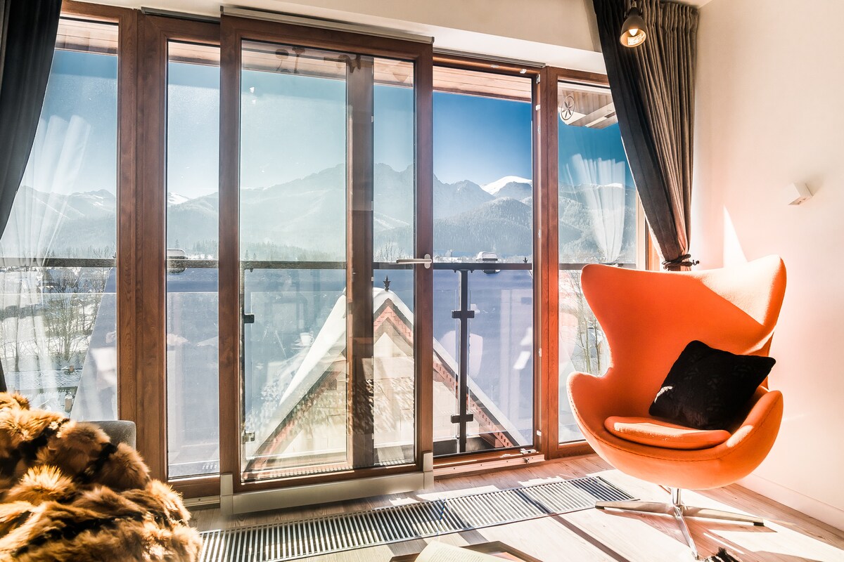 Large glass doors open to a stunning view of mountains and water, inviting natural light into the space. A modern orange chair anchors the corner, while a cozy fur throw lies on the floor, enhancing the inviting interior.