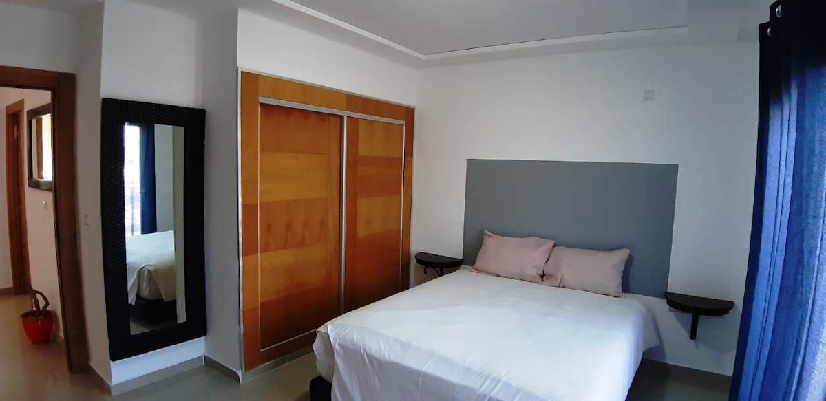 The bedroom features a spacious bed with light-colored bedding. A wall-mounted headboard is set against a neutral backdrop. Two small side tables frame the bed, while a full-length mirror stands adjacent to a sliding closet with wooden accents. Natural light enters through the window covered by dark curtains.