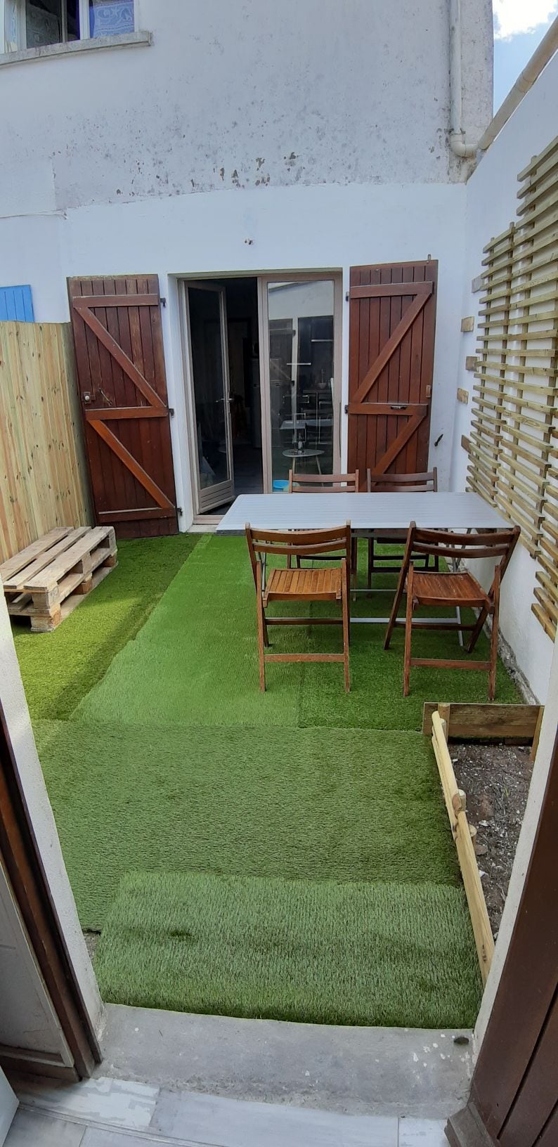 A small courtyard features artificial grass with two wooden chairs positioned around a simple table. Natural light is visible through the open double doors leading into the interior space. Wooden fencing surrounds the area, contributing to a sense of privacy.
