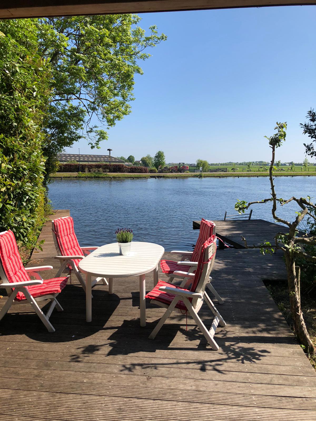 An outdoor seating area is featured by the water, with four white chairs and a round table positioned under a bright blue sky. A small decorative plant sits on the table, while the tranquil river and lush greenery create a serene environment.