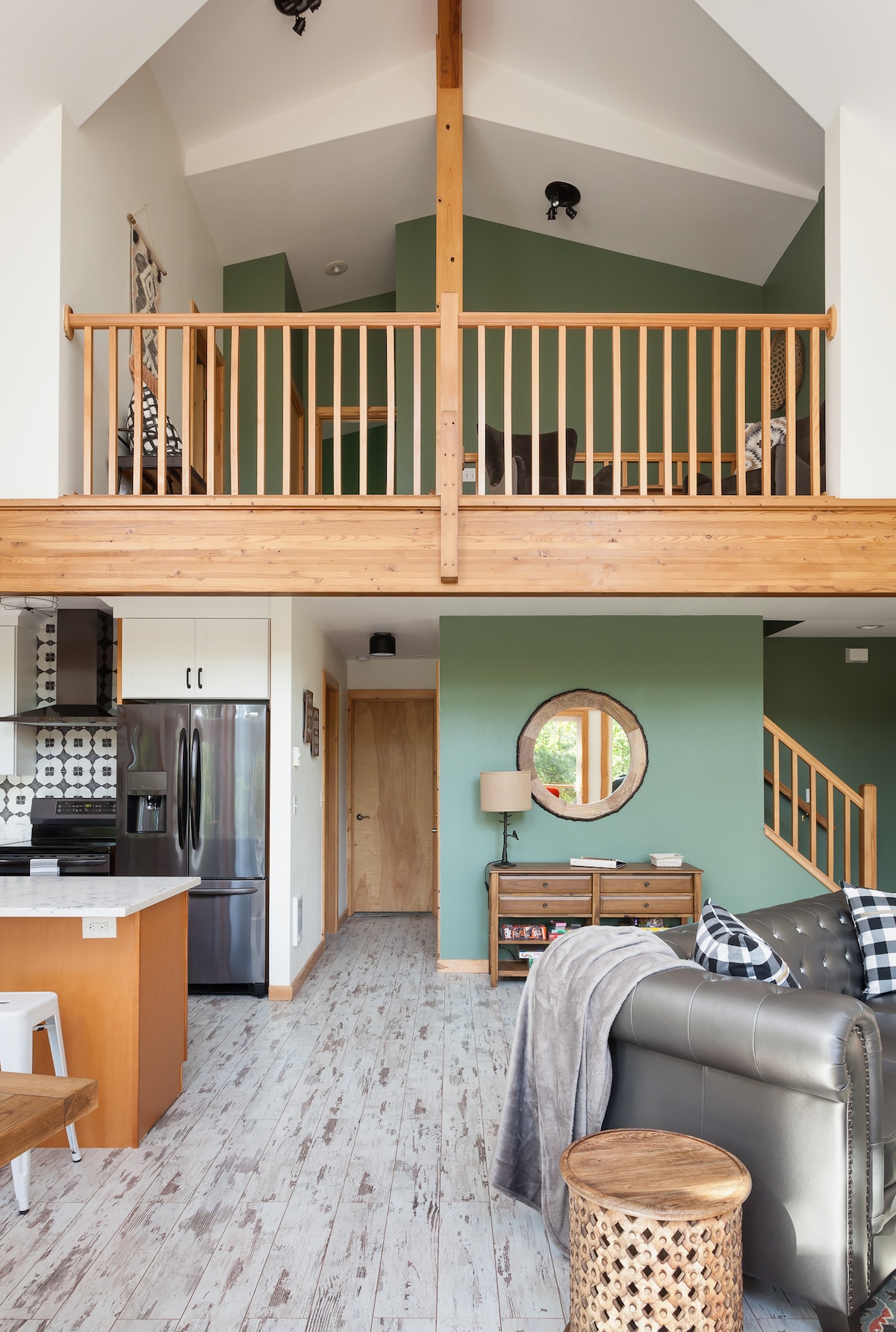 An open-concept living area features a modern kitchen with stainless steel appliances and wooden accents. A staircase leads to a loft space, with a railing and seating visible. The walls are painted in a calming green tone, complemented by natural light filtering through large windows.