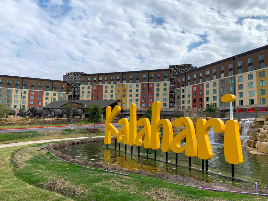A vibrant yellow sign displaying 'Kalahari' is positioned in front of a large resort building. The resort features a mix of colorful facades and large windows. A landscaped area with a small pond and stone features is visible in the foreground, offering a sense of openness.