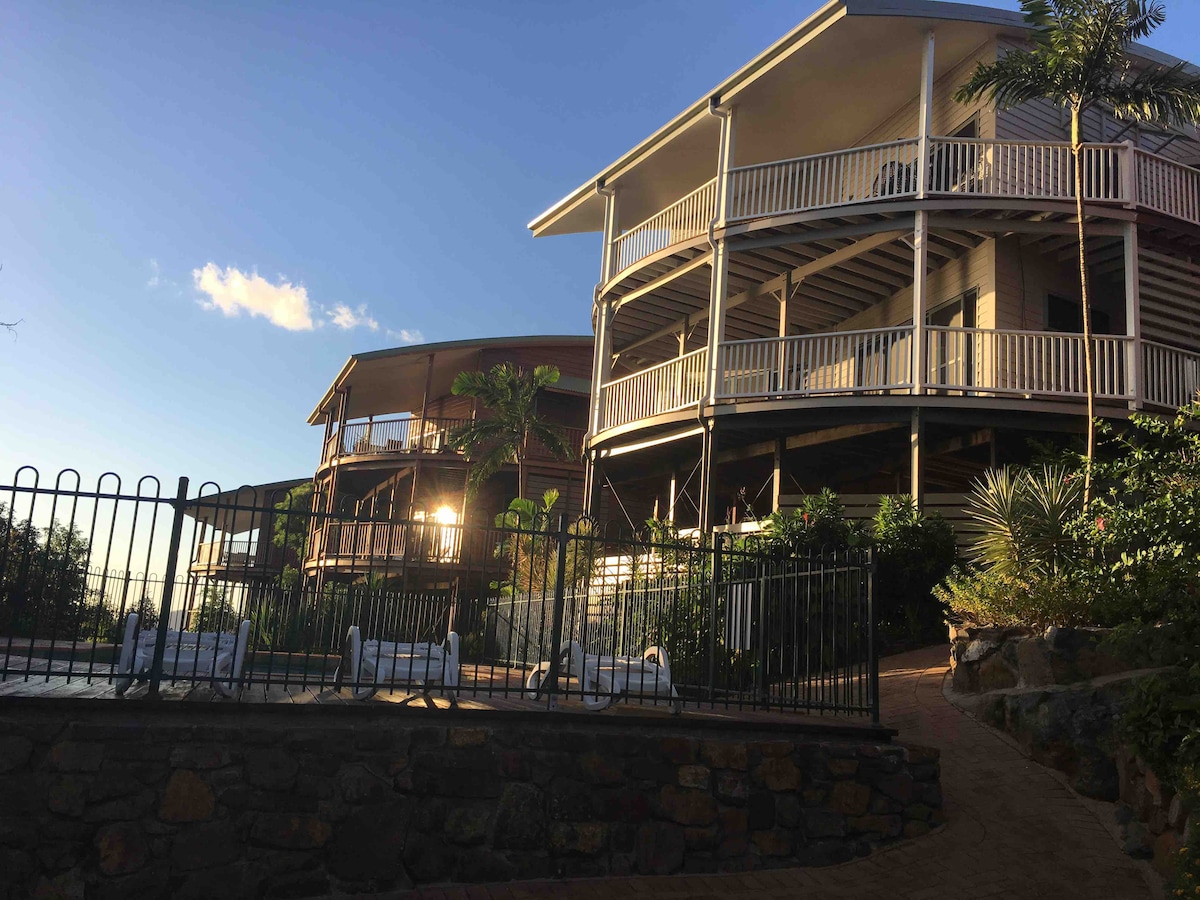 The villa features an upper and lower level with expansive balconies, surrounded by lush greenery. A stone pathway leads to the property, with a fence defining the perimeter. Soft sunlight casts a warm glow on the structure, accentuating the architectural details.