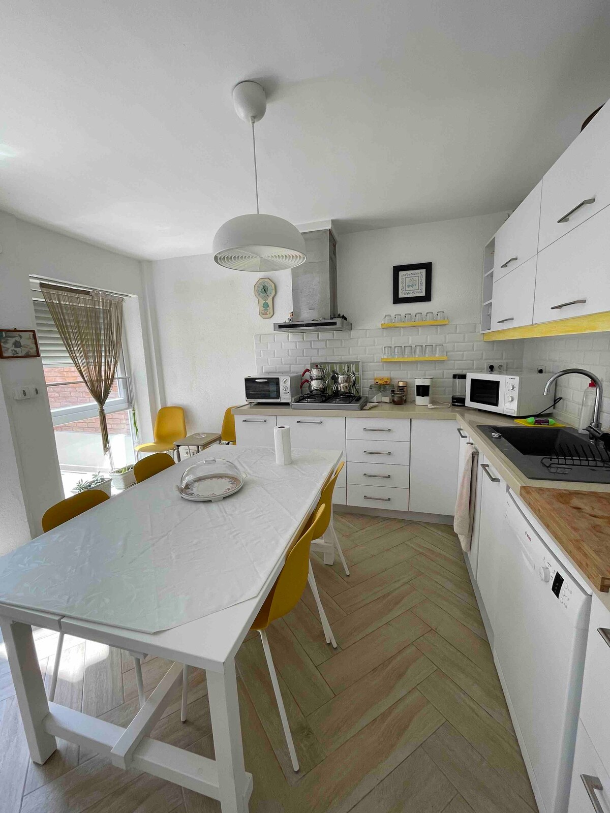 A newly equipped kitchen features white cabinetry and modern appliances, including a microwave and stove. A spacious table is surrounded by yellow chairs, while natural light filters through the window, enhancing the bright and functional atmosphere.