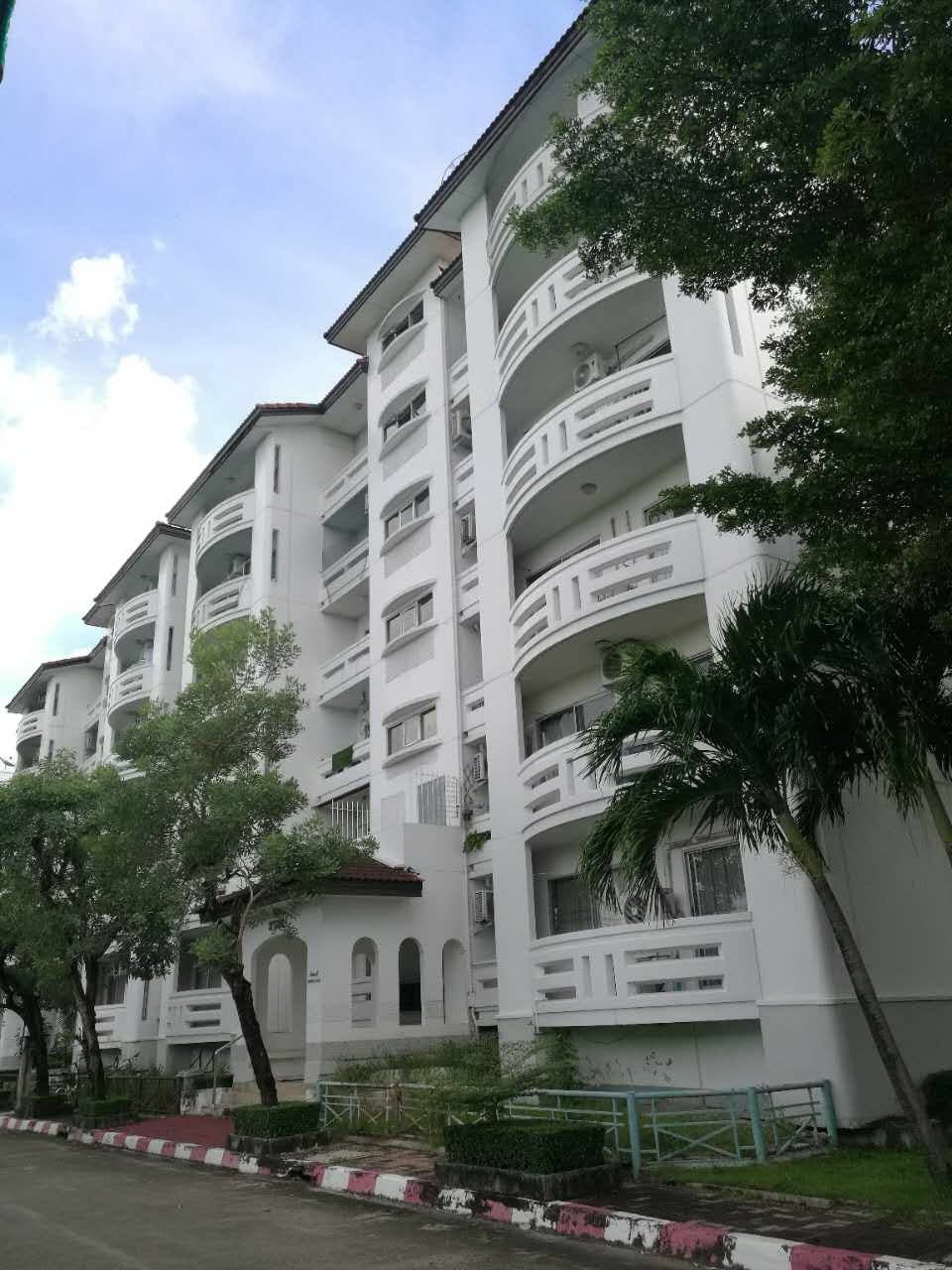 A multi-story white condominium building is shown, featuring multiple balconies with decorative railings. Lush green trees are positioned in front, and the sky above is bright with scattered clouds, contributing to a vibrant outdoor environment.
