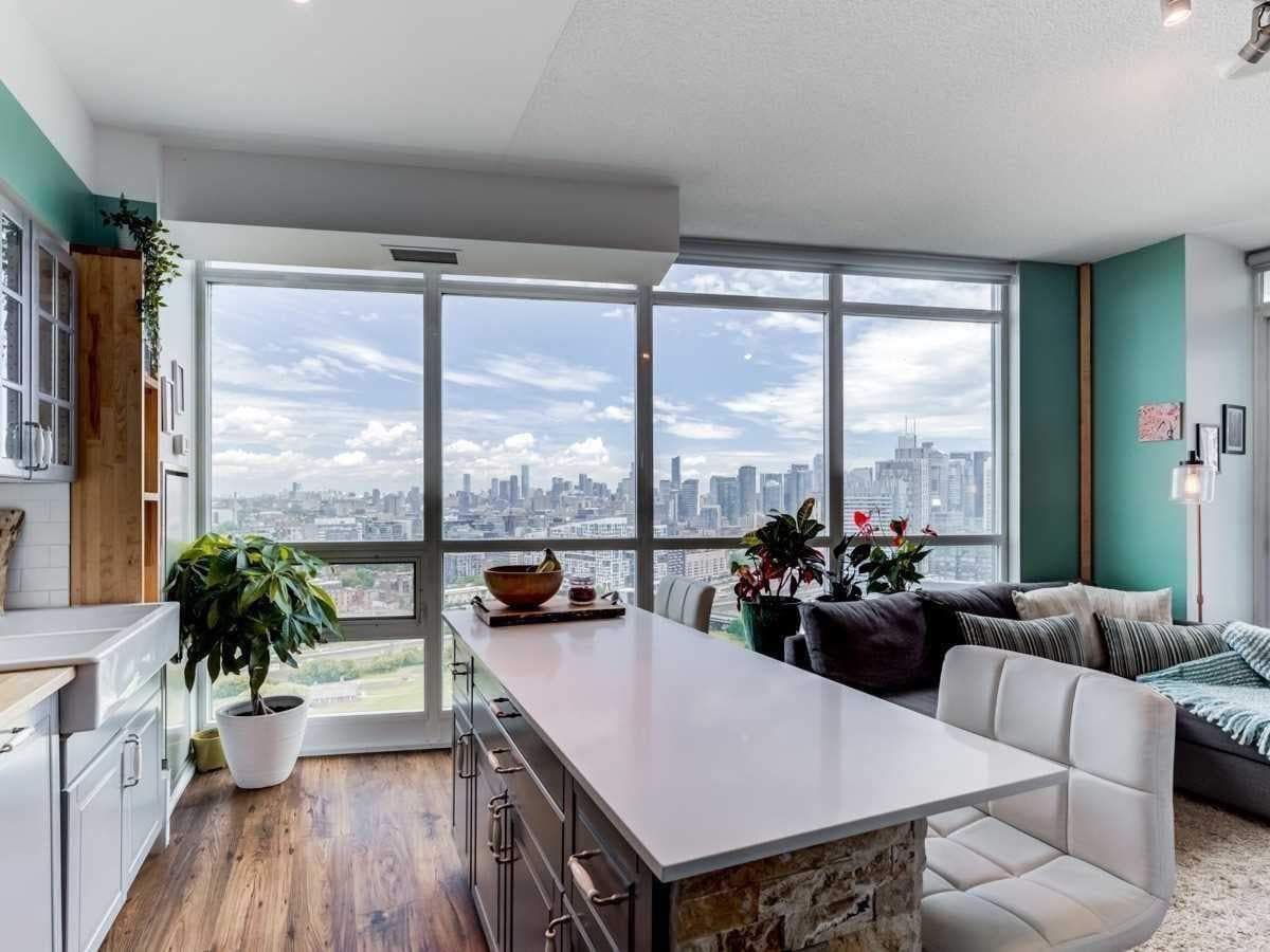 A spacious living area features large windows offering panoramic city views. Bright green accent walls complement a modern kitchen with a long island. A cozy seating area is positioned to enjoy the skyline, along with potted plants adding a touch of nature.
