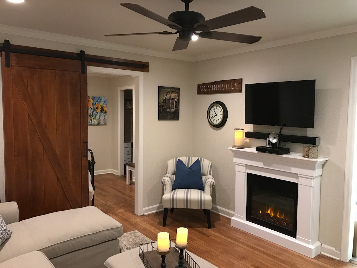 The living room features a comfortable seating area with a light-colored couch, complemented by a striped armchair. A modern electric fireplace is situated beneath a wall-mounted television, while decorative elements, such as a wall clock and artwork, enhance the cozy ambiance.