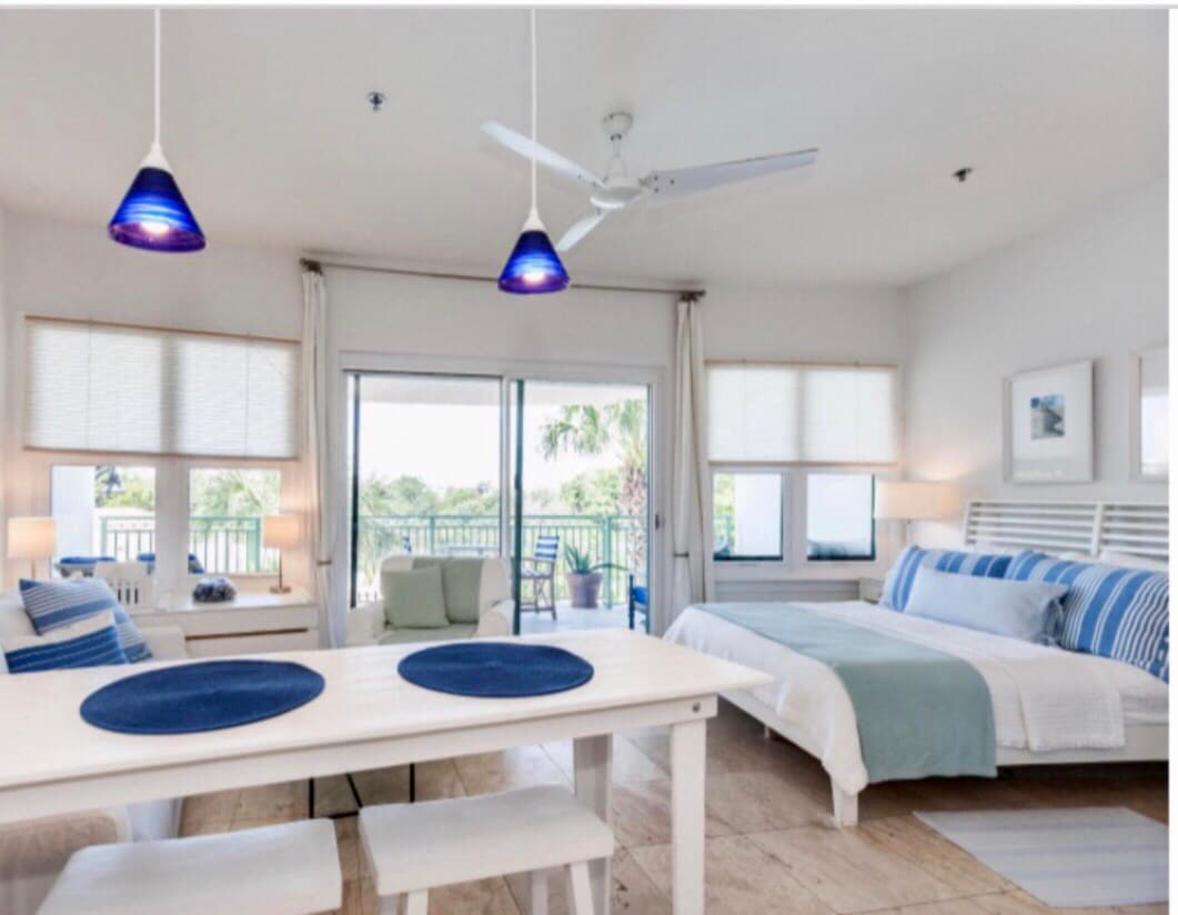 An inviting studio space features a spacious bed dressed in light linens, flanked by subtle blue accents. A dining table is set with blue placemats, and large windows allow natural light to fill the room. A ceiling fan and pendant lights add functionality and modernity.