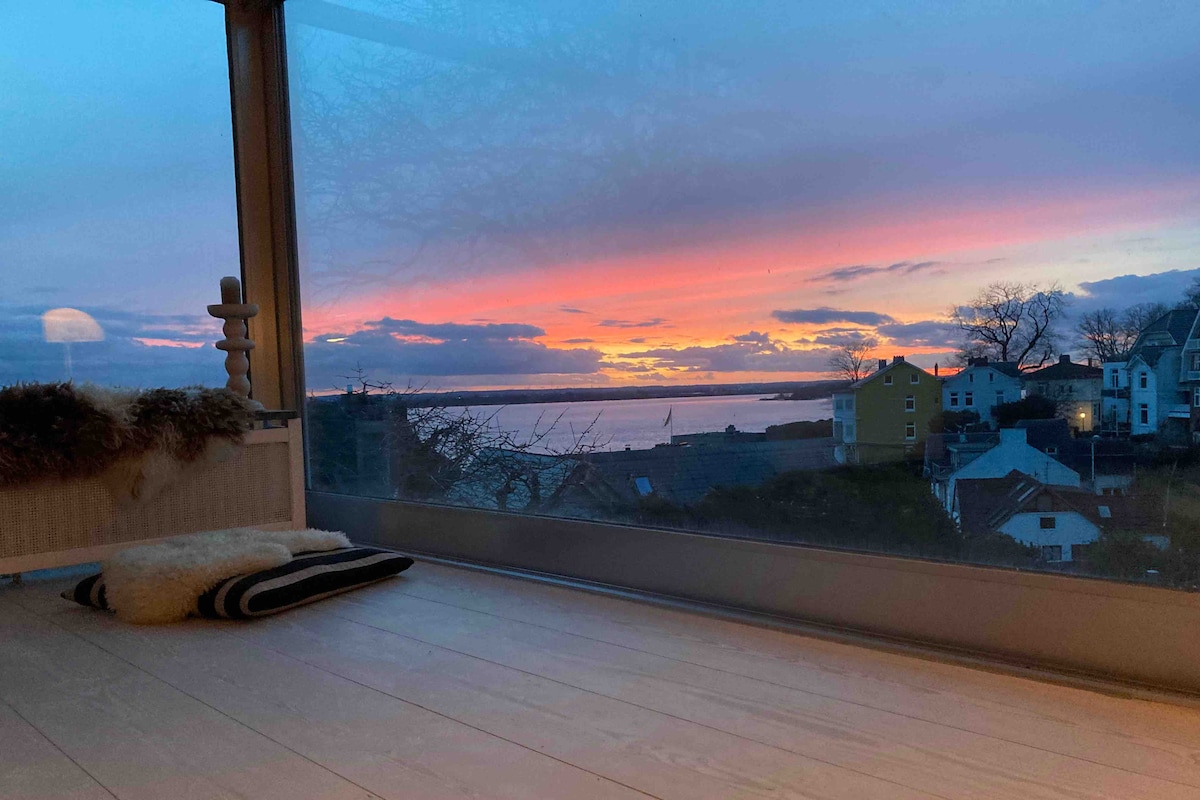 A large window offers a view of a vibrant sunset over the water, with hues of orange, pink, and purple reflected in the sky. The space appears to have a cozy area with a soft rug positioned on light wooden flooring.