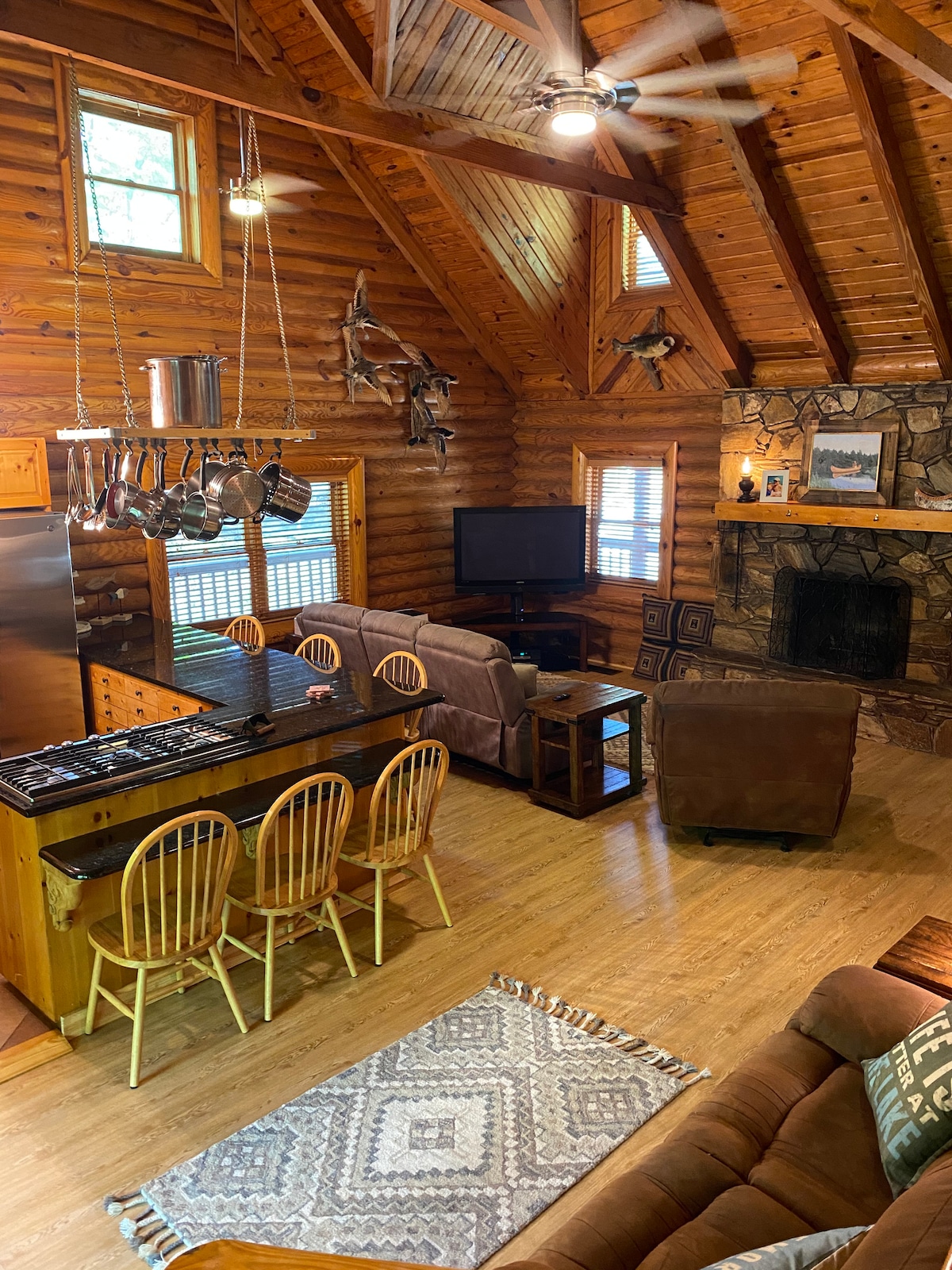 An open-concept living area features a log cabin design with high ceilings and wooden beams. A kitchen with a bar seating four is visible, alongside a cozy sitting area. Natural light enters through multiple windows, illuminating the warm tones of the furnishings.