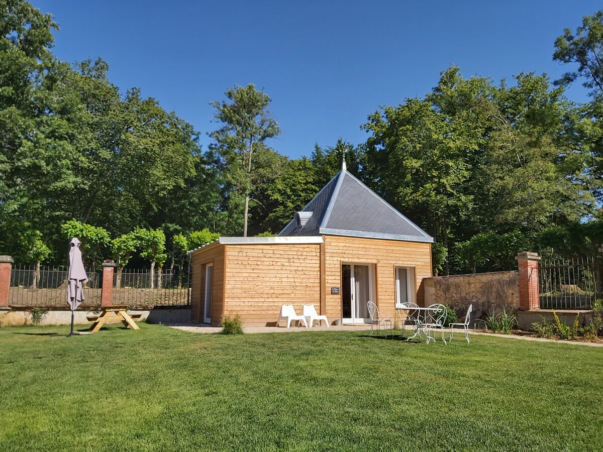 A charming gîte is set against a backdrop of lush greenery, featuring a wooden exterior and a distinctive peaked roof. Outdoor seating arrangements are visible on the manicured lawn, complemented by trees that provide shade and a serene atmosphere.