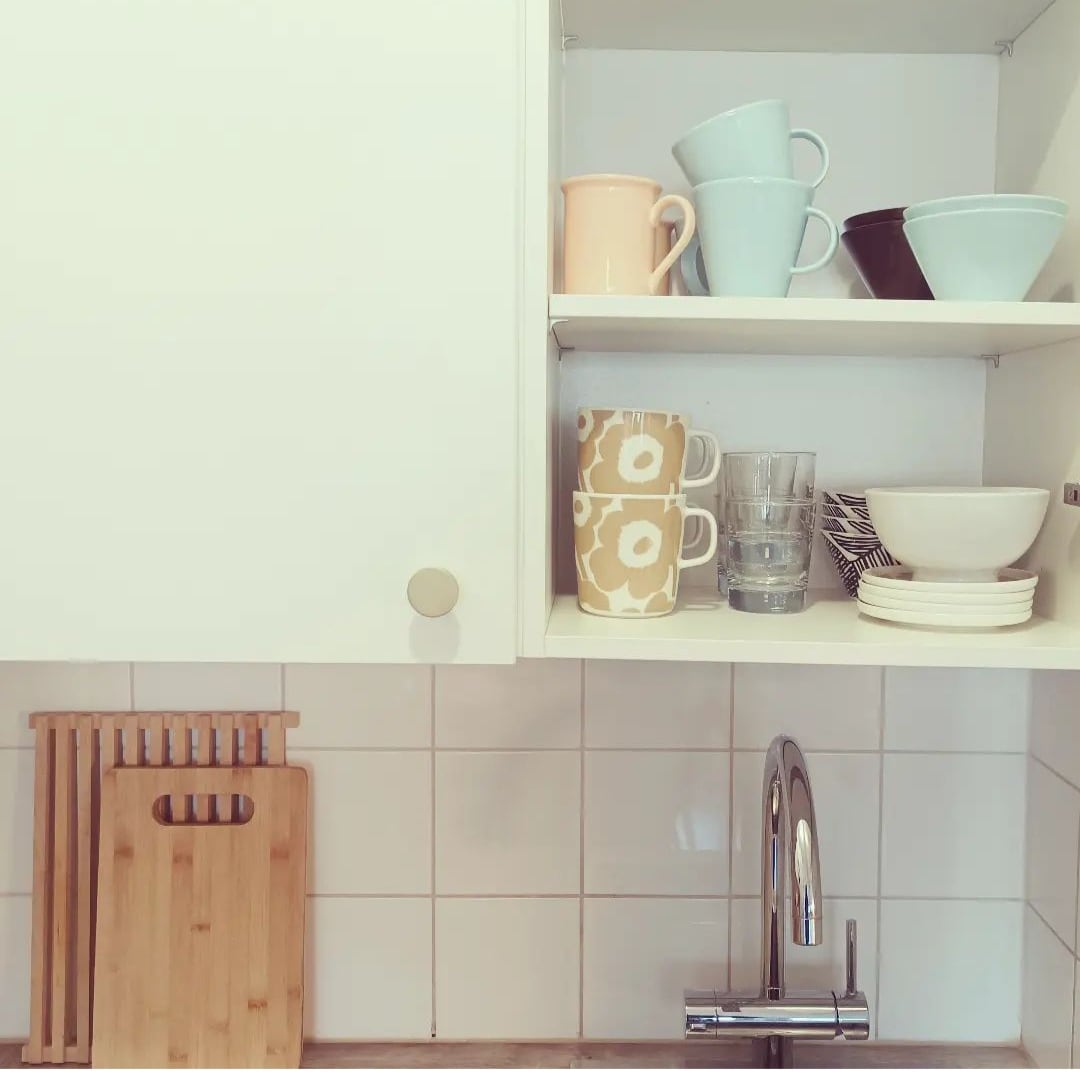 A well-equipped open kitchen is displayed, featuring neatly arranged dishware on white shelves. Various cups, bowls, and glasses are visible, alongside a wooden cutting board and kitchen utensils. The sink area features a modern faucet, blending functionality with simplicity.