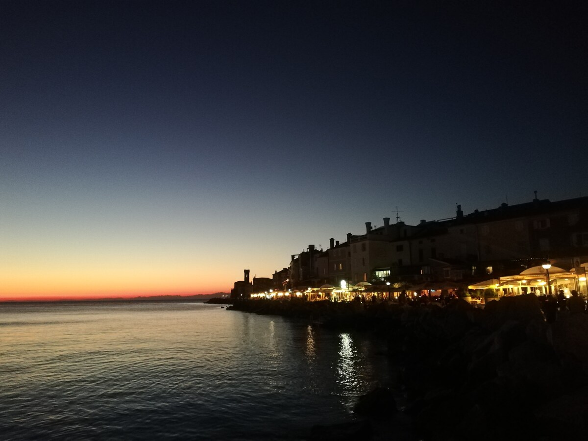 The image captures a serene coastal view during twilight, where the horizon displays a gradual transition from deep blue to warm orange tones. Illuminated buildings line the waterfront, reflecting softly on the calm water. The atmosphere appears peaceful and inviting.