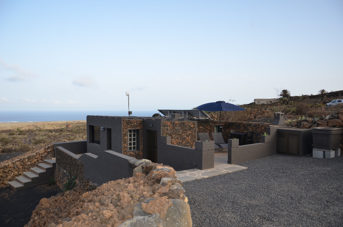 The rustic house blends with its natural surroundings, featuring stone walls and a contemporary touch. Outdoor seating areas are visible, alongside a large umbrella and the gravel driveway leading to the property. The ocean can be seen in the distance, enhancing the serene landscape.