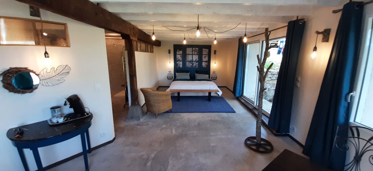 An open and airy room features a spacious bed against a backdrop of blue accent walls, complemented by artwork. A comfortable chair is positioned near a small table with a coffee setup. Large glass doors invite natural light, leading to an outdoor area.