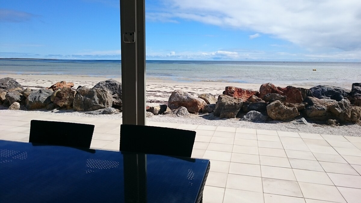A view of the beachfront features a clear blue sky and calm waters. The foreground includes large, smooth rocks along the shore, while a patio table with chairs is seen, providing an inviting space for outdoor dining.