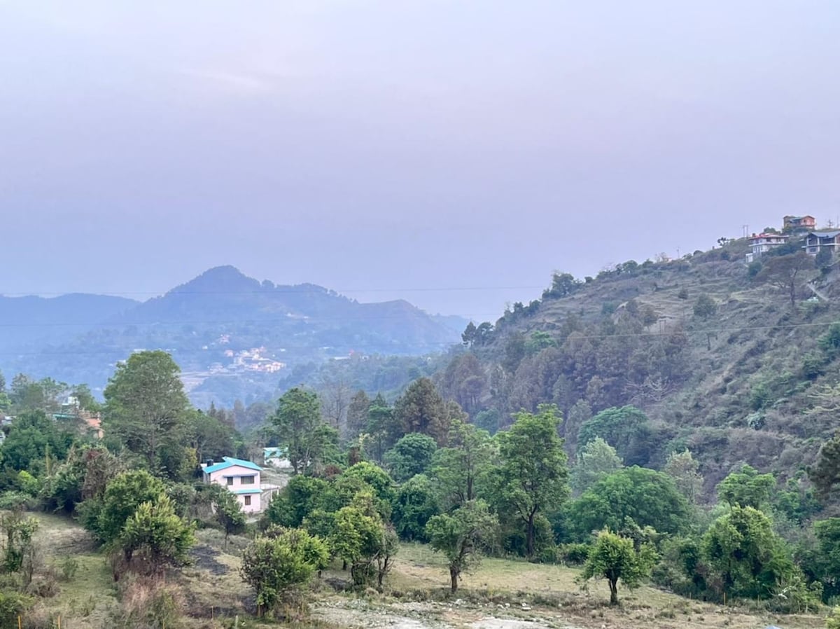A scenic view features rolling hills covered in greenery, dotted with clusters of trees and a few distant homes. The soft hues of the sky blend harmoniously, creating a tranquil backdrop for the natural landscape.