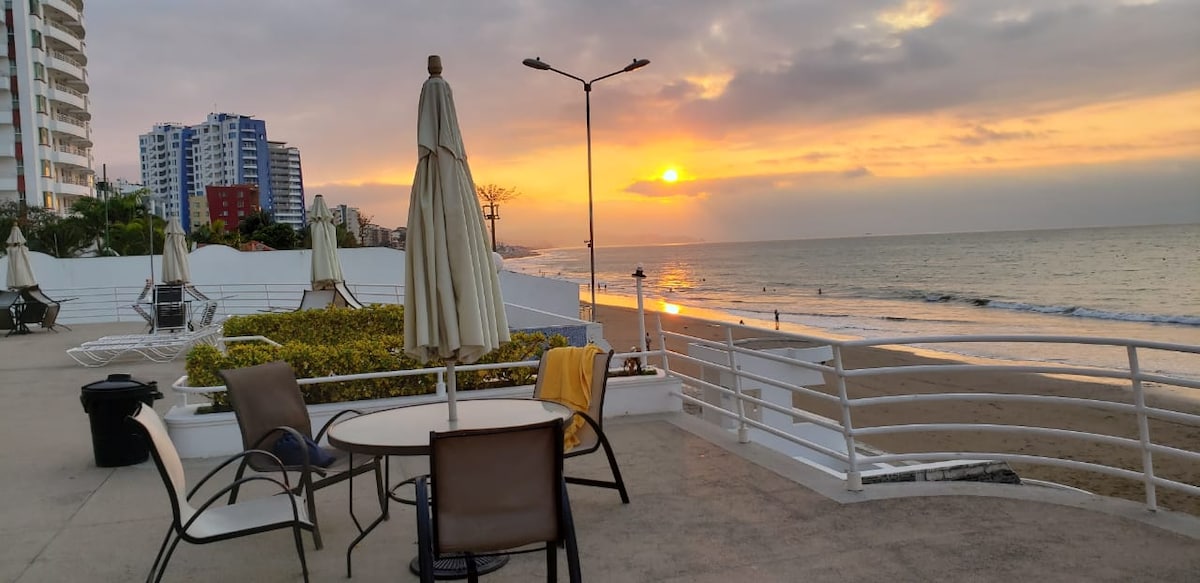 The image captures a serene beachside view during sunset, with gentle waves lapping at the shore. A round table and chairs are positioned on a terrace, accompanied by a large umbrella. The horizon displays a warm sunset glow, while the coastal buildings are visible in the background.