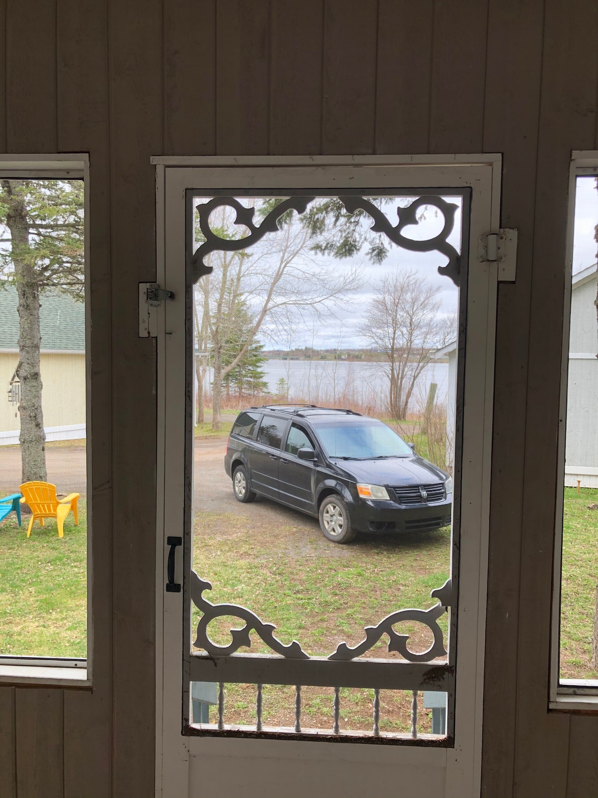 The entryway features a decorative screen door, providing a clear view of a parked vehicle in the driveway. Outside, a grassy area leads to the calm waters of the bay, framed by trees. Colorful outdoor chairs are visible nearby, enhancing the inviting setting.