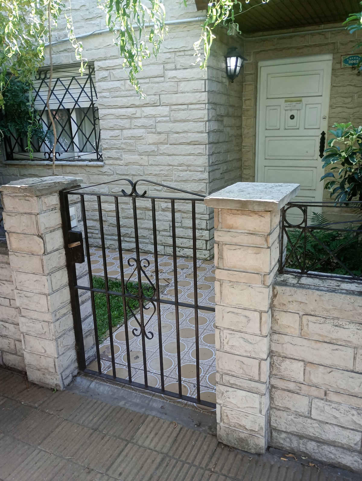 An entrance is framed by a decorative metal gate that opens onto a tiled pathway leading to the house. The stone exterior wall of the property and a well-kept garden with greenery are visible, creating an inviting approach.
