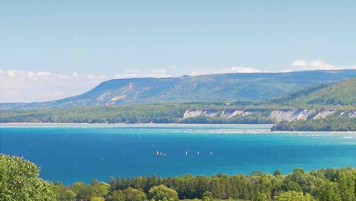 The expansive view captures the tranquil waters of Georgian Bay, framed by lush greenery and rugged hills. The clear blue sky complements the vibrant turquoise hues of the water, while distant sailboats add a sense of activity to the serene landscape.