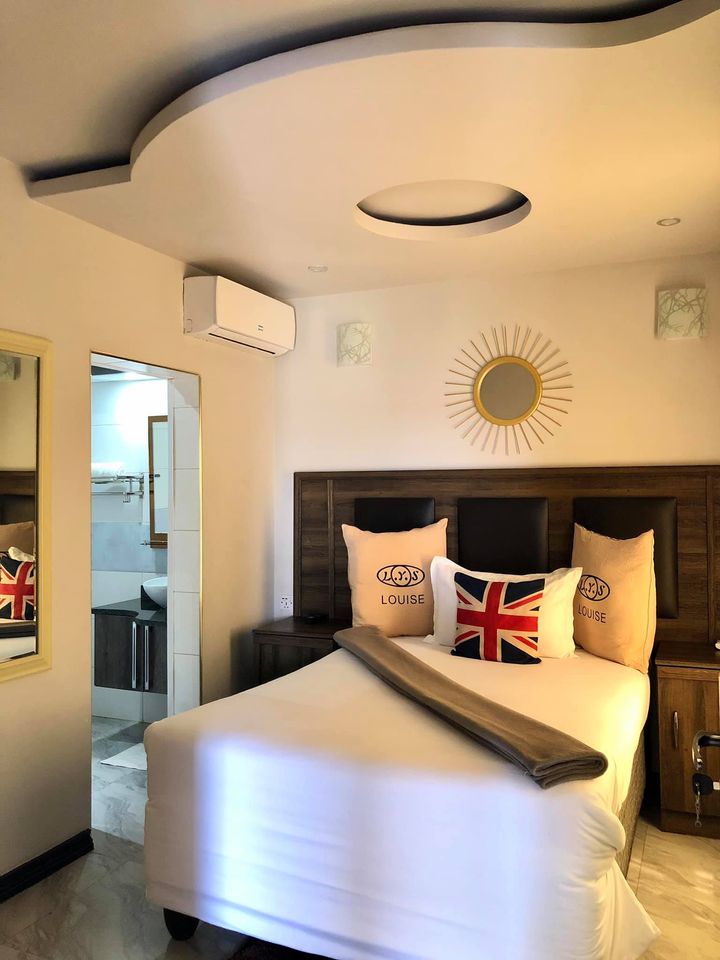 A cozy room features a spacious bed adorned with soft pillows and a decorative blanket. A mirror reflects light, enhancing the room's brightness. A circular ceiling detail adds interest, while air conditioning and soft lighting contribute to a comfortable atmosphere.