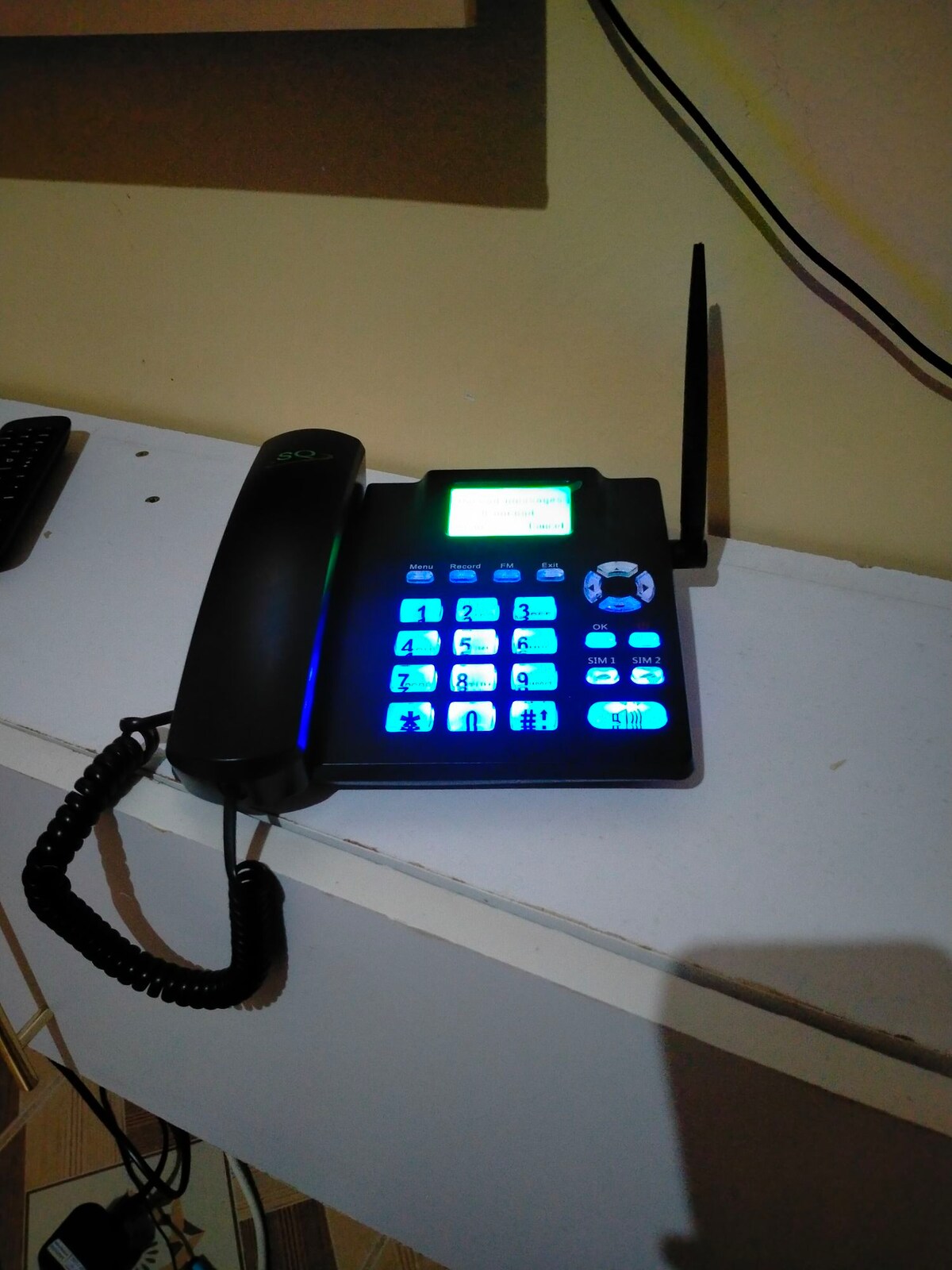 A black desktop phone with a coiled handset is positioned on a light-colored surface. The phone features a keypad with illuminated blue buttons and a small digital display, showcasing various function options. Nearby, wires are visible against the wall.