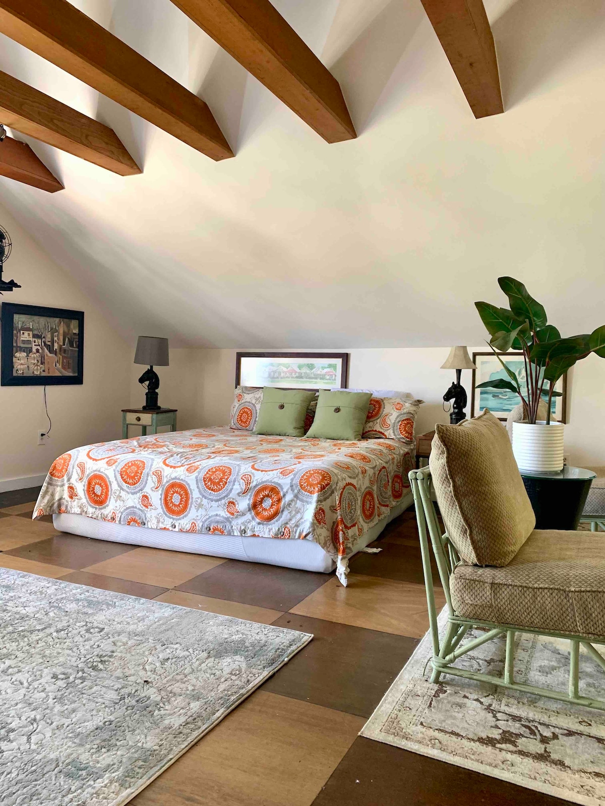 A spacious bedroom features a king-sized bed adorned with a patterned comforter and coordinating pillows. Natural light illuminates the room, highlighting the wood beams on the ceiling. A seating area with an armchair and side table complements the decor, while potted plants add a touch of greenery.