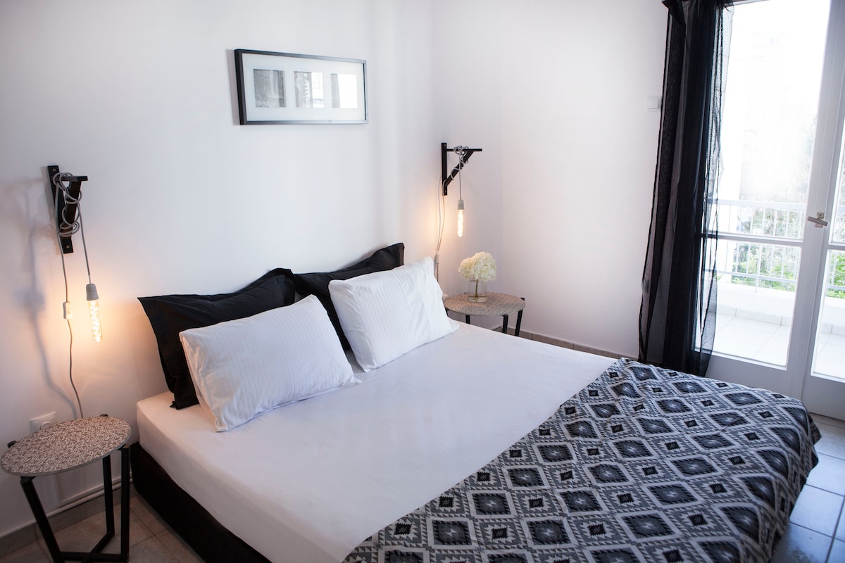 A well-lit sleeping area features a spacious bed adorned with white linens and a patterned blanket. Two bedside tables with stylish lamps are positioned on either side. A sliding glass door leads to a balcony, providing natural light and a view of the outdoors.