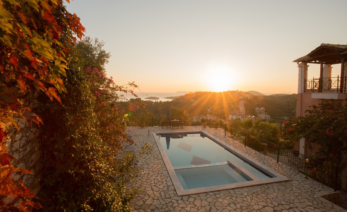 A serene outdoor area features a swimming pool framed by stone tiles, with vibrant foliage nearby. The sun sets in the distance, casting a warm glow over the landscape, while hills and distant islands are visible on the horizon.