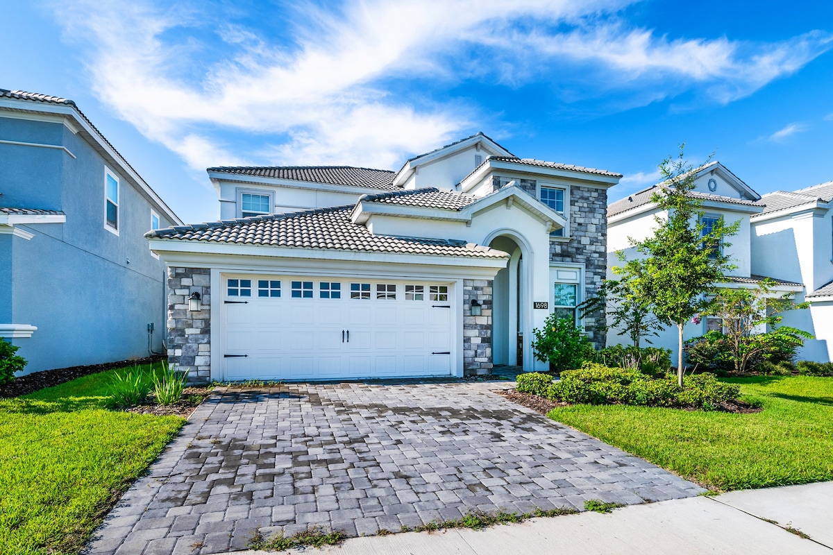 A modern villa exterior is showcased with a stone and stucco facade. The driveway, constructed of pavers, leads to a double garage with white doors. Lush green landscaping, including small trees and shrubs, complements the home against a clear blue sky.
