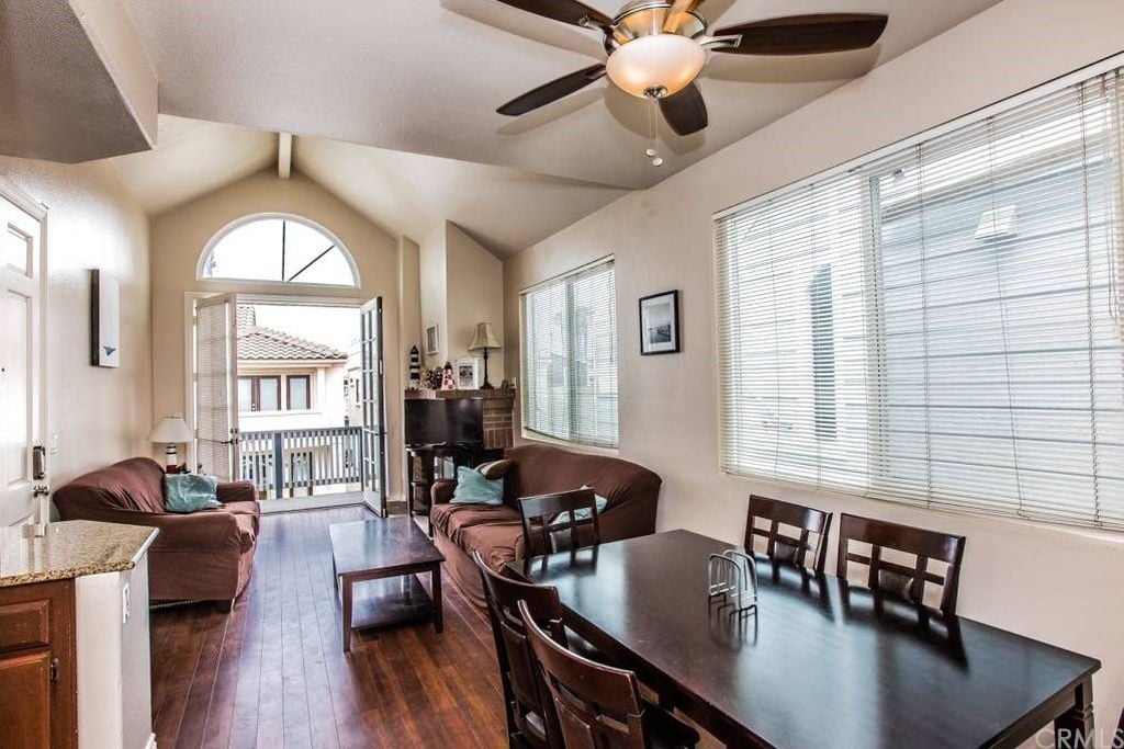 A spacious living area features large windows with natural light flowing in, creating a bright atmosphere. Comfortable seating arrangements are provided with two sofas and a dining table surrounded by chairs. A ceiling fan adds to the airy feel, while an open door leads to the outdoor space.
