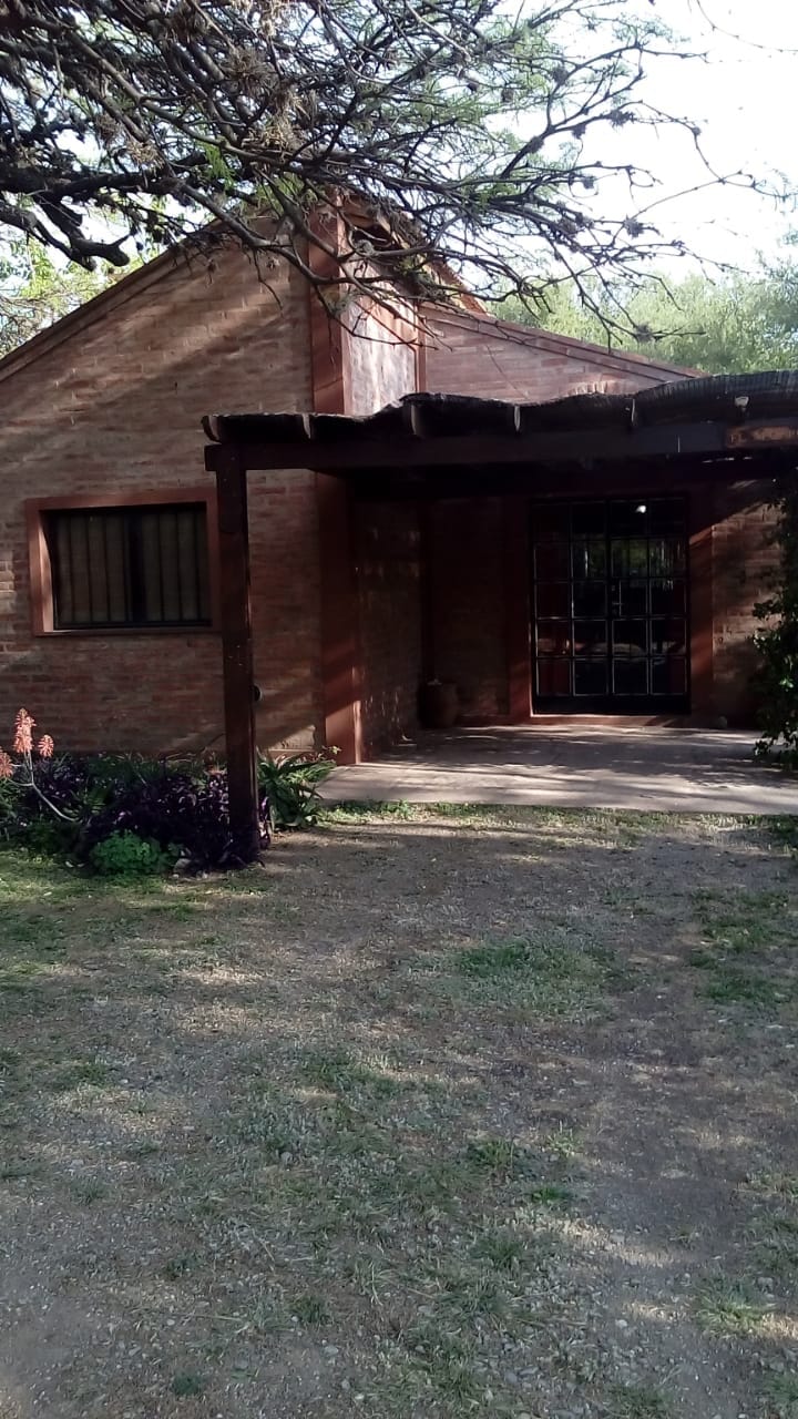 The exterior of the house is displayed, featuring a wooden pergola over the entrance. Surrounding greenery and a gravel pathway lead to the door, with brick walls providing a rustic charm. A small garden with blooming plants adds color to the serene setting.