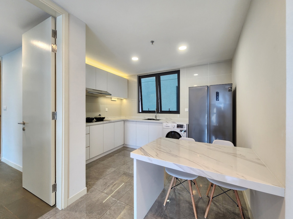 A modern kitchen is equipped with sleek cabinetry and essential appliances, including a stove and microwave. A compact dining area features a minimalistic table paired with four chairs. Natural light enters through a large window, illuminating the space.
