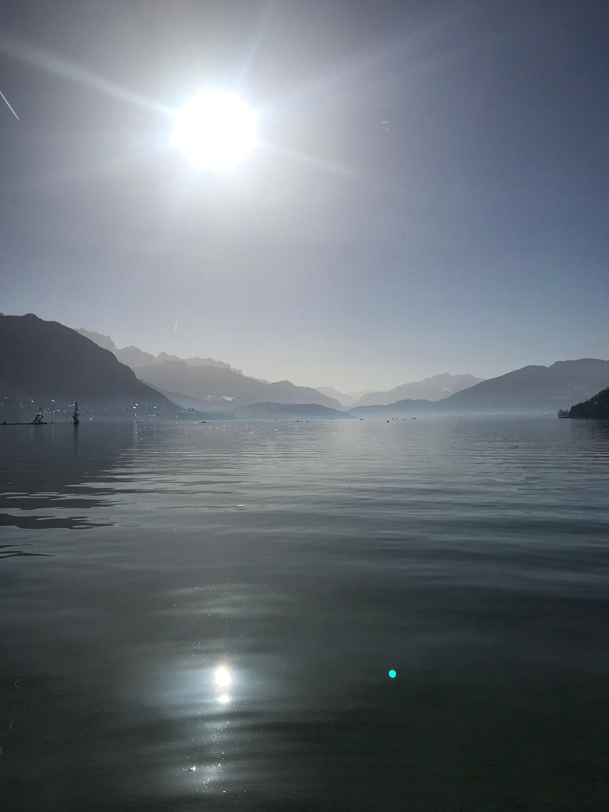 A serene lake scene is captured, reflecting the mountains and sky. The sun emits a bright glow over the tranquil water, creating a calm atmosphere. Silhouettes of distant peaks are visible against the horizon, and the surface of the lake appears smooth and glass-like.