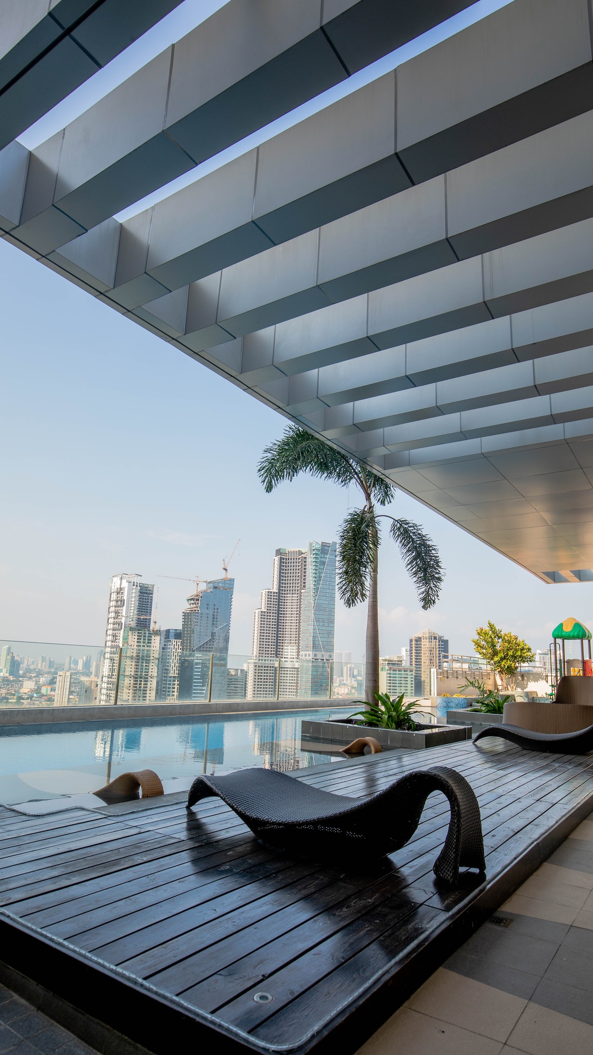 A serene rooftop pool area features wooden decking with modern lounge chairs positioned beside a glistening infinity pool. Towering skyscrapers can be seen in the distance, framed by clear blue skies and hints of greenery from potted plants.
