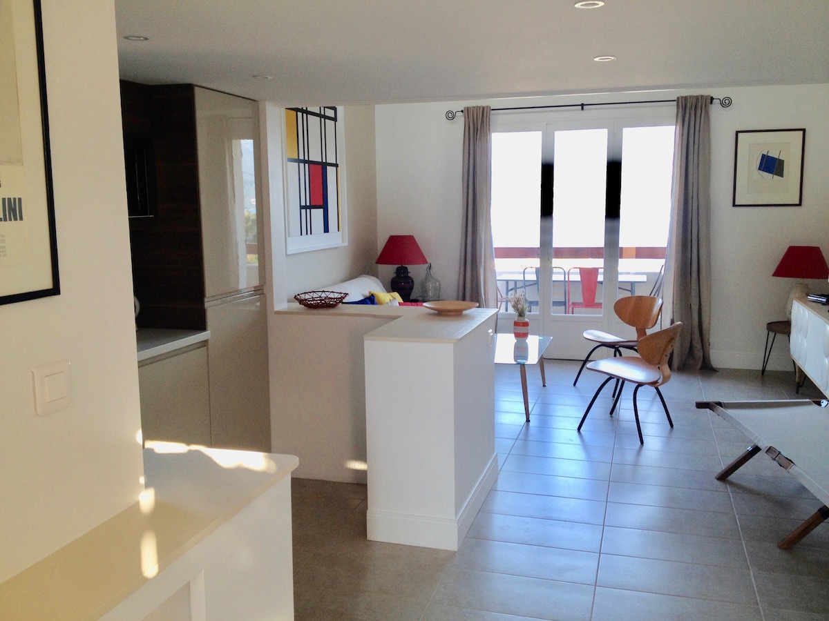 An open living area features modern decor, with a light color palette and natural light flooding in through large glass doors. A comfortable chair is positioned next to a small table, while the kitchen showcases sleek cabinetry. A beautiful view of the outdoor terrace is visible.
