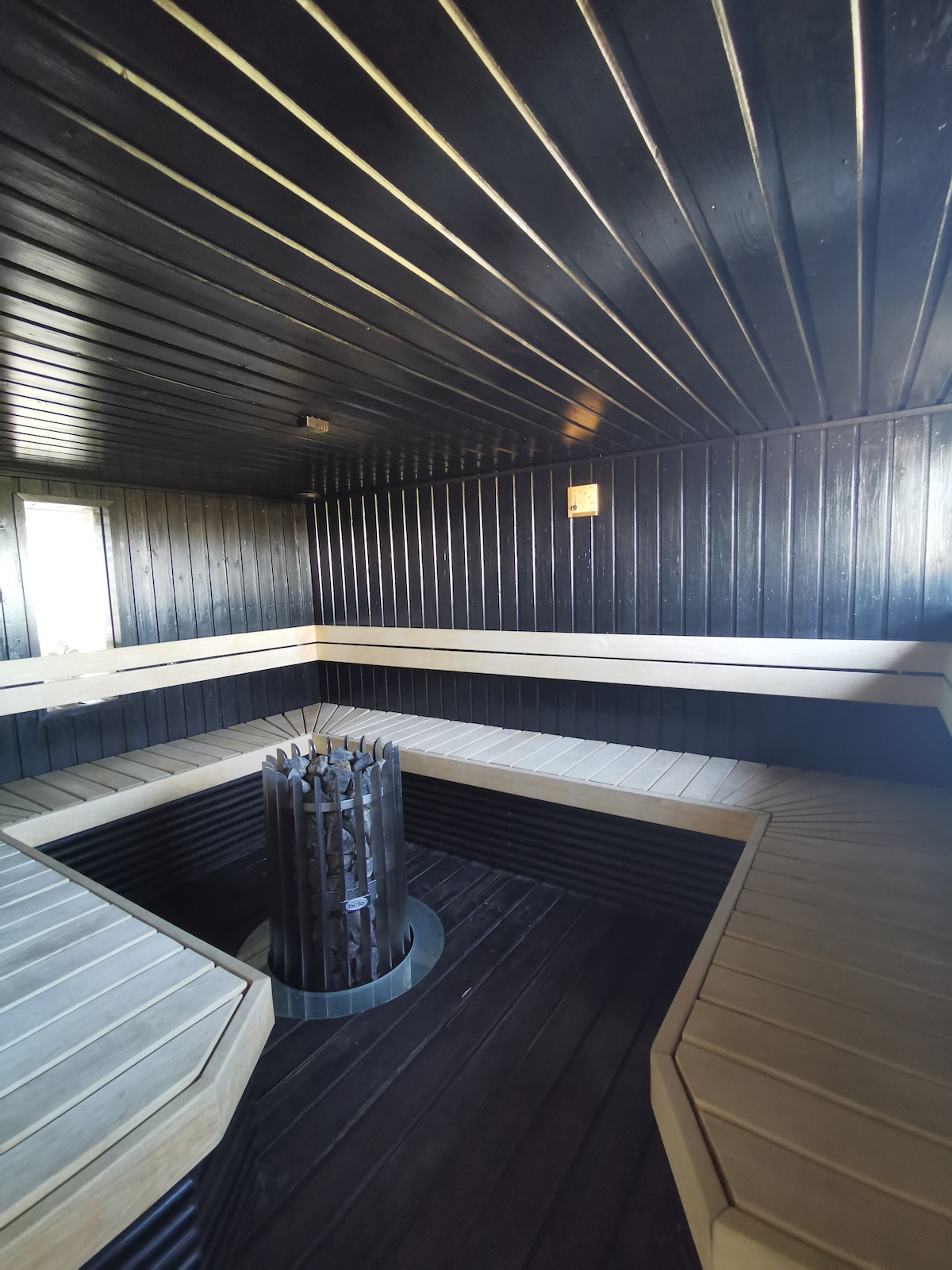 The sauna features dark wooden paneling with a ceiling of slatted wood. Wooden benches encircle a central stove, contributing to a relaxing atmosphere. Windows provide natural light, allowing for a bright and airy feel while maintaining privacy.
