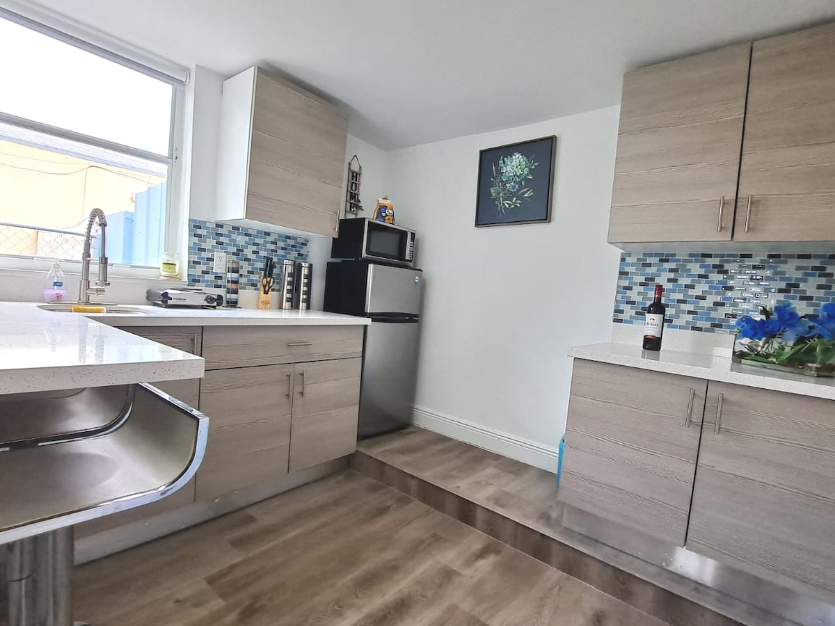 A functional kitchen area features modern cabinets, a stainless steel refrigerator, and a sleek countertop. A blue-tiled backsplash adds a pop of color. Natural light streams in from a nearby window, highlighting the space's clean lines and practical layout.