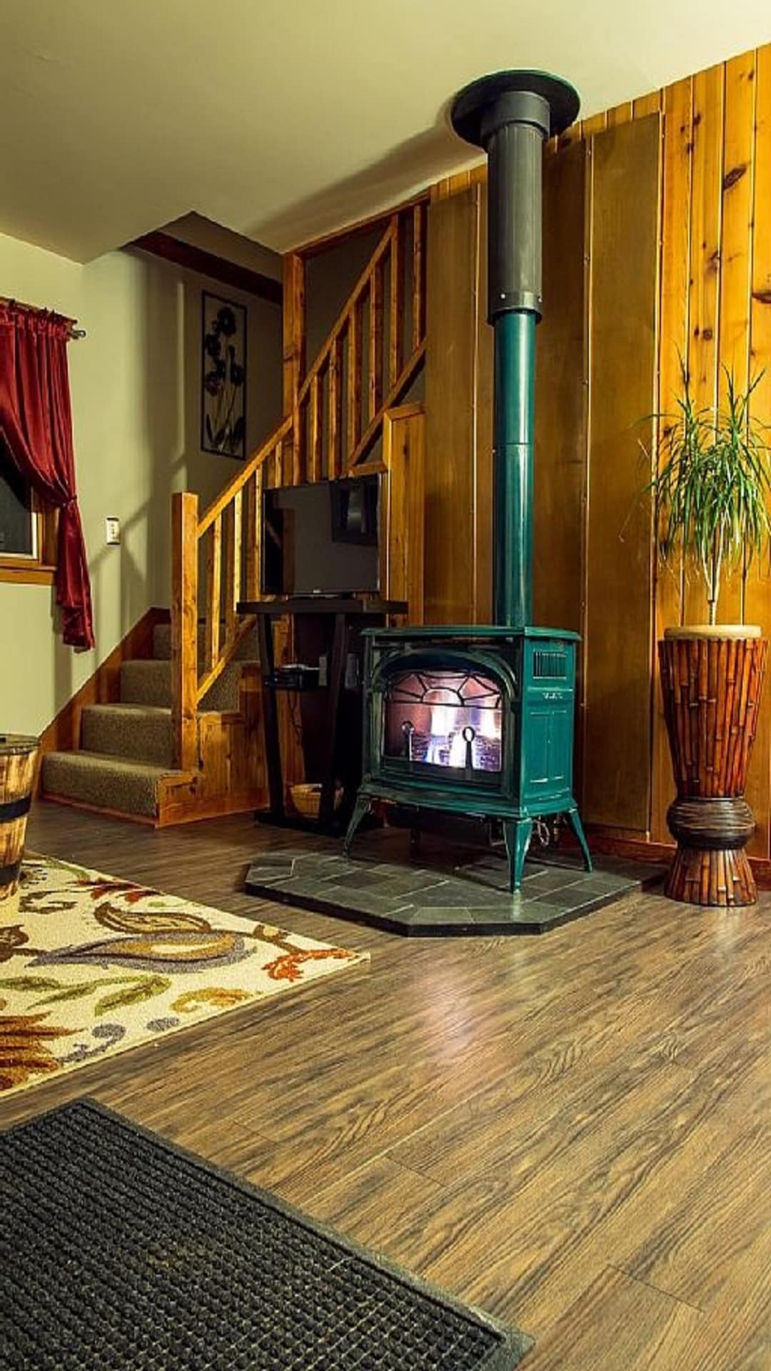 A wood-burning stove in a cozy corner adds warmth to the living space. The surrounding walls feature a mixture of wood paneling and soft paint. A staircase leads to the upper level, while a decorative plant and patterned rug enhance the welcoming environment.