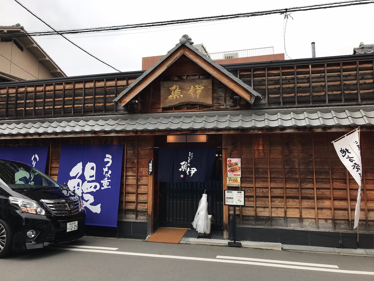 The exterior of a traditional Japanese building is featured, showcasing wooden paneling and a tiled roof. A welcoming entrance is marked by a wooden sign above. Decorative banners and a modern vehicle are visible in front, enhancing the blend of tradition and contemporary life.