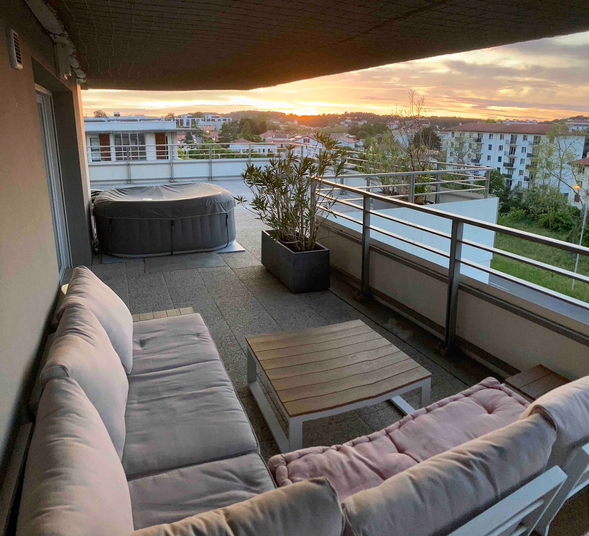 A spacious rooftop terrace is equipped with a comfortable seating area and a wooden coffee table. A hot tub is partially visible, and greenery is present in a large planter. The sunset casts a warm glow over the surrounding landscape and rooftops.