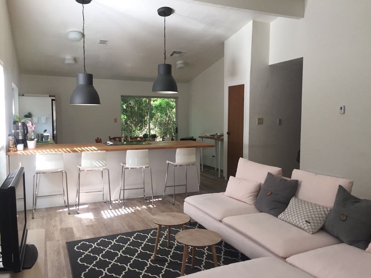 An open living area features a light-colored sectional sofa, accompanied by two small wooden tables. A kitchen island with bar stools provides a casual dining space. Large windows allow natural light to filter in, framing views of the surrounding greenery.