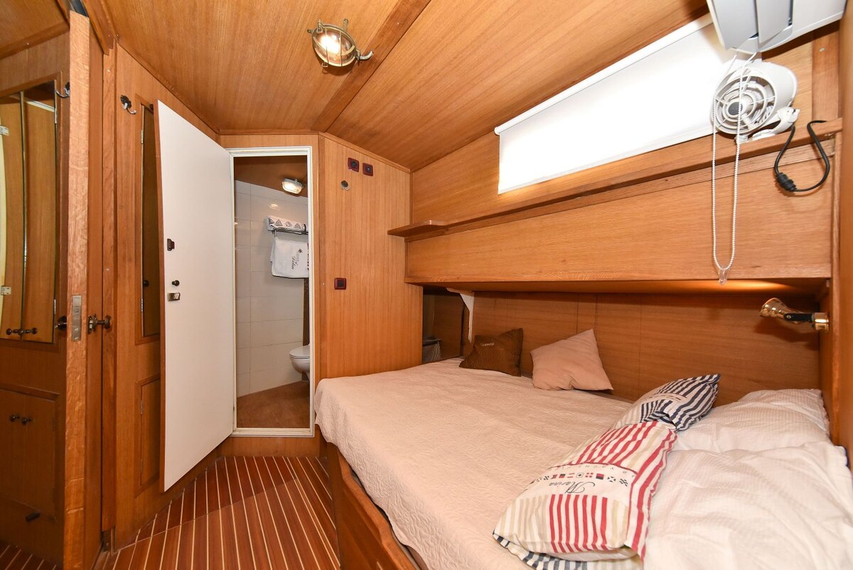 A warm wood-paneled cabin features a cozy sleeping area with a spacious bed, adorned with light-colored pillows. A door leads to an adjoining bathroom, while air conditioning controls are visible on the wall. Soft light filters through a small window above the bed.